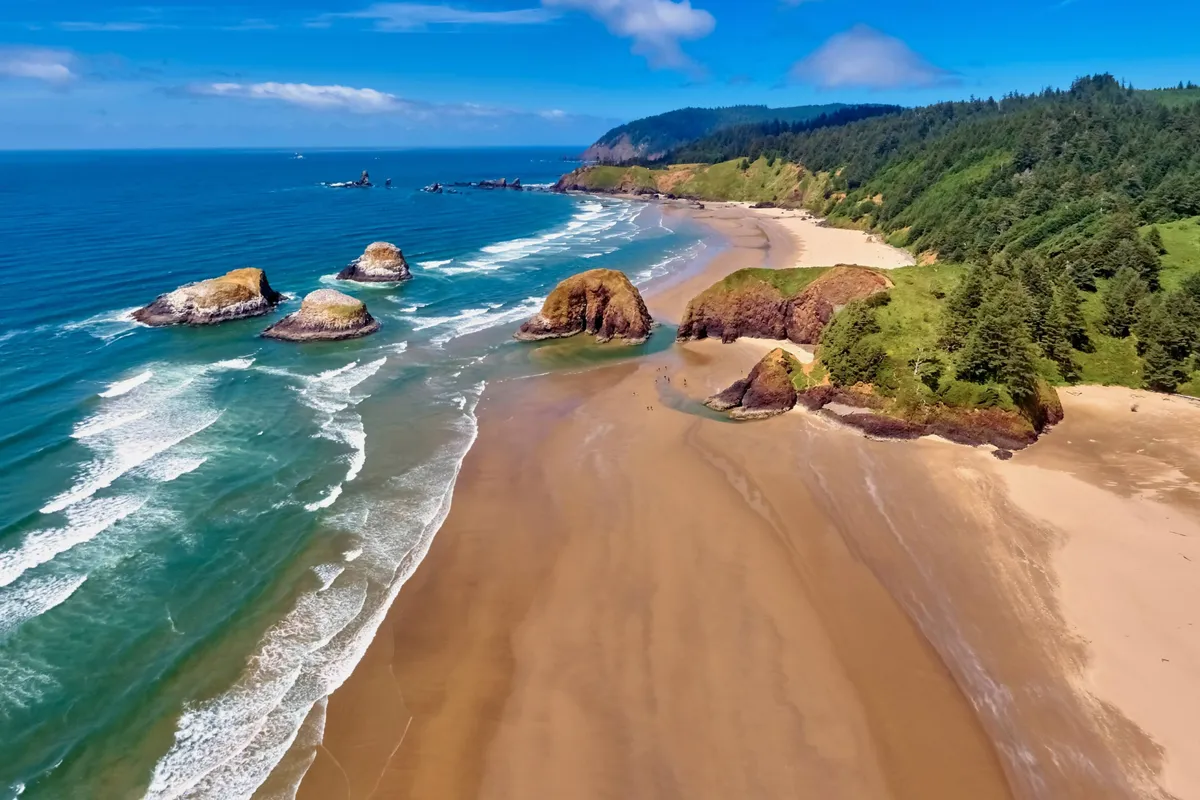 A sweeping exterior shot of the Oregon Coast, featuring expansive sand beaches, rolling ocean waves, striking rock formations, and green hills in the background. The scene is serene and inviting, perfectly capturing the natural beauty of the Rockaway Beach area.