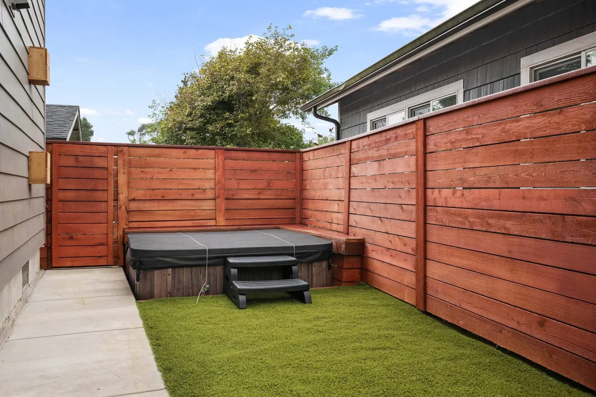 A tranquil backyard scene featuring a hot tub set on artificial grass, surrounded by a wooden fence for privacy. The setting suggests peaceful relaxation and opportunities for group gatherings under the open sky.