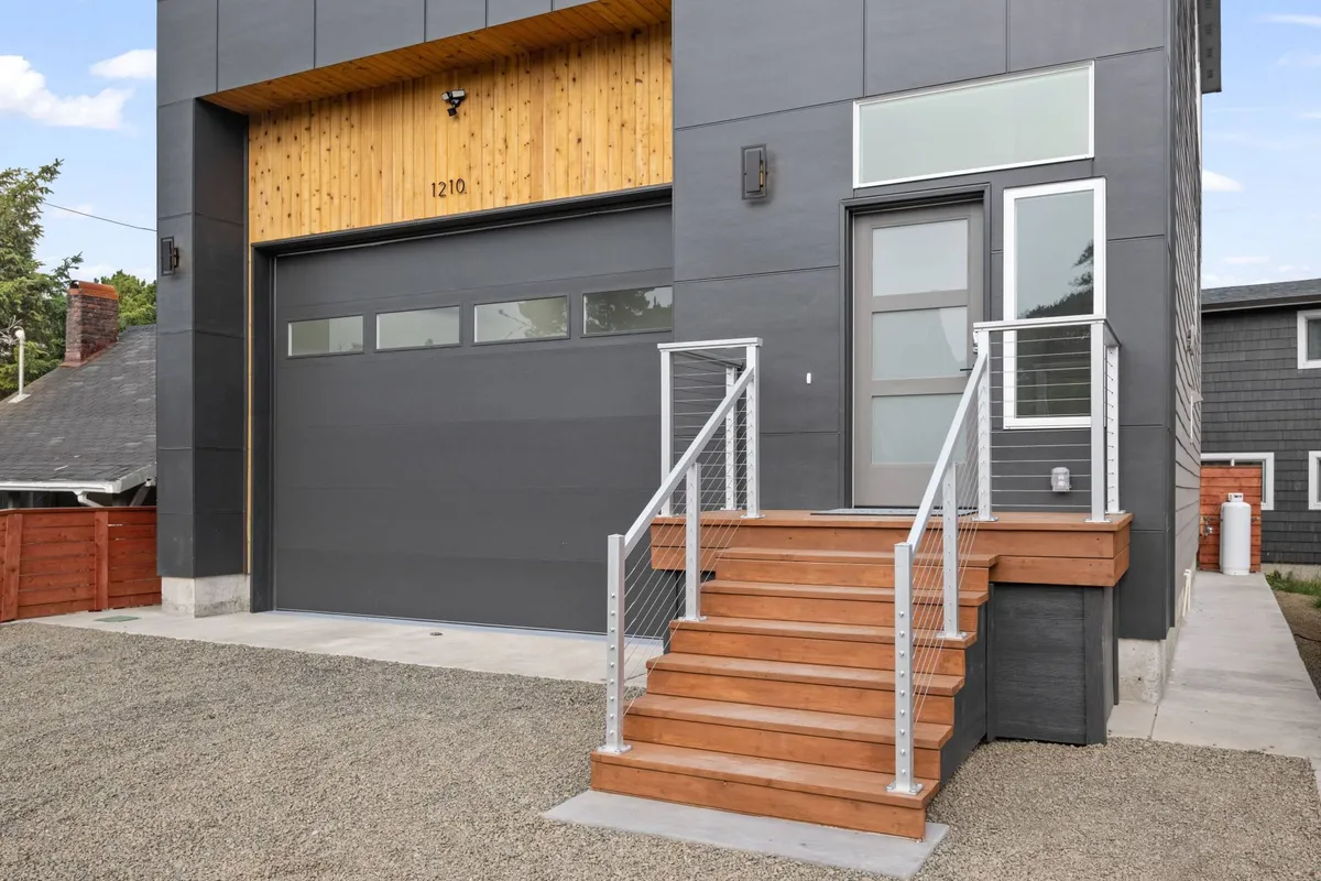 A welcoming modern exterior featuring wooden stairs, a garage door, a sleek front door, and a metal railing. The front area offers visible parking and convenient access points.