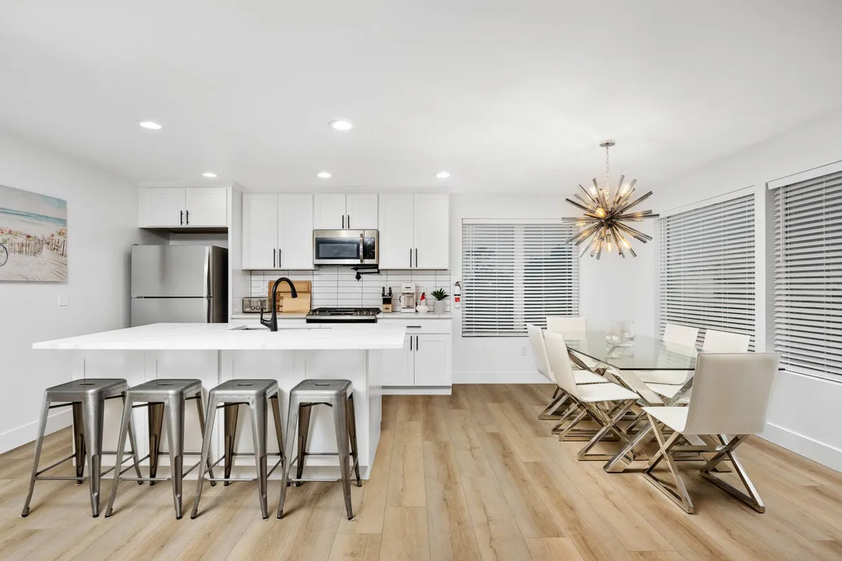 A modern dining area with an island and bar stools adjacent to a spacious dining table surrounded by stylish chairs. Decorative lighting enhances the contemporary ambiance, and the open layout encourages gathering for meals or social events.
