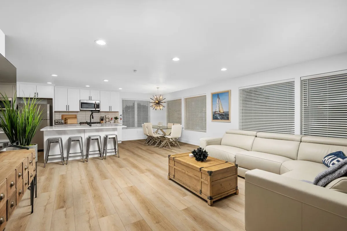 The image displays an open-concept living area with a kitchen island, dining table, a comfortable sofa, and colorful artwork on the wall. The layout feels inviting and spacious, ideal for gathering and relaxing after a day of beach or trail adventures.