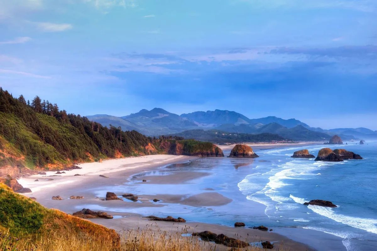 A wide, inviting sand beach stretches along the coastline with ocean waves rolling in, dramatic rock formations in the distance, and lush green hills and forests beyond. The scene is tranquil with soft light, suggesting an ideal setting for beachcombing, playing in the sand, or family walks by the water.