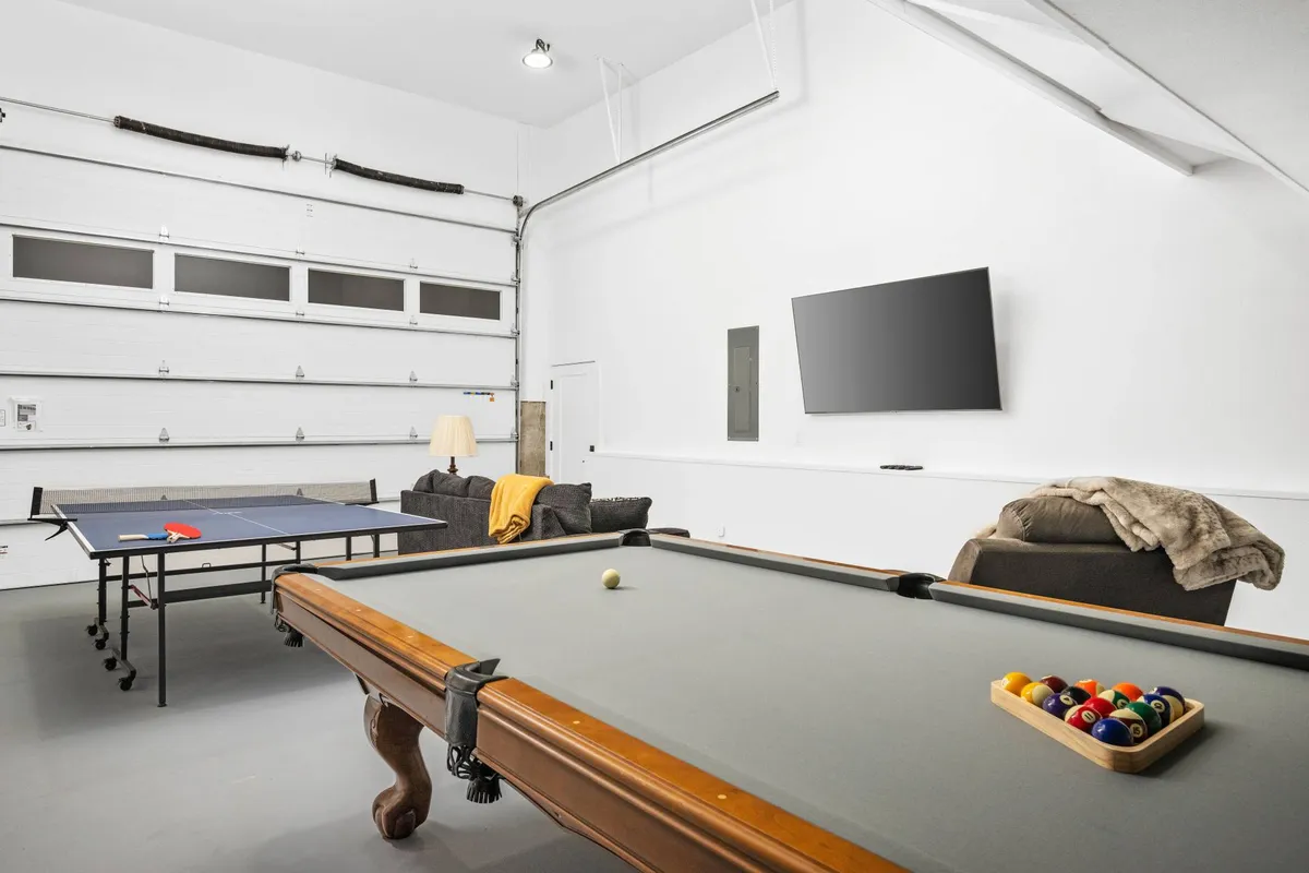 A spacious, inviting game room featuring a pool table and a ping pong table as centerpieces. A large, comfortable sofa is arranged facing a wall with a sizable television, and the room is bathed in natural light. The space is clearly designed for group entertainment and relaxation, capturing an atmosphere of playful competition and communal enjoyment.