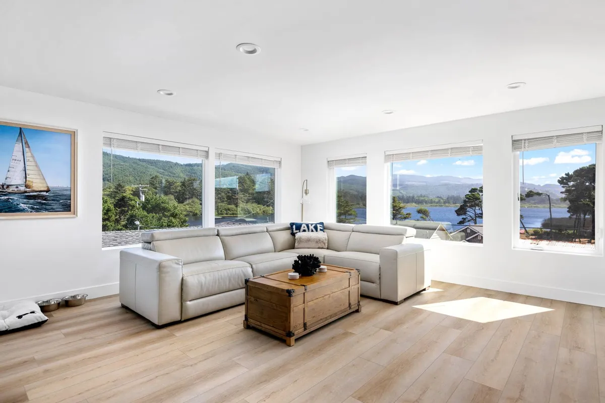 A spacious living area featuring a large sectional sofa and a wooden coffee table, set against expansive windows that showcase breathtaking lake views. Landscape artwork adds a touch of serenity, and the abundance of natural light creates an inviting, tranquil atmosphere perfect for relaxation or gathering with loved ones.