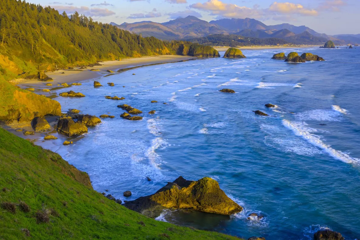 A rugged yet beautiful stretch of coastline with waves crashing on rocks, a sandy beach in the foreground, and distant mountains under a glowing sky. The scene exudes tranquility and invites exploration and outdoor play.
