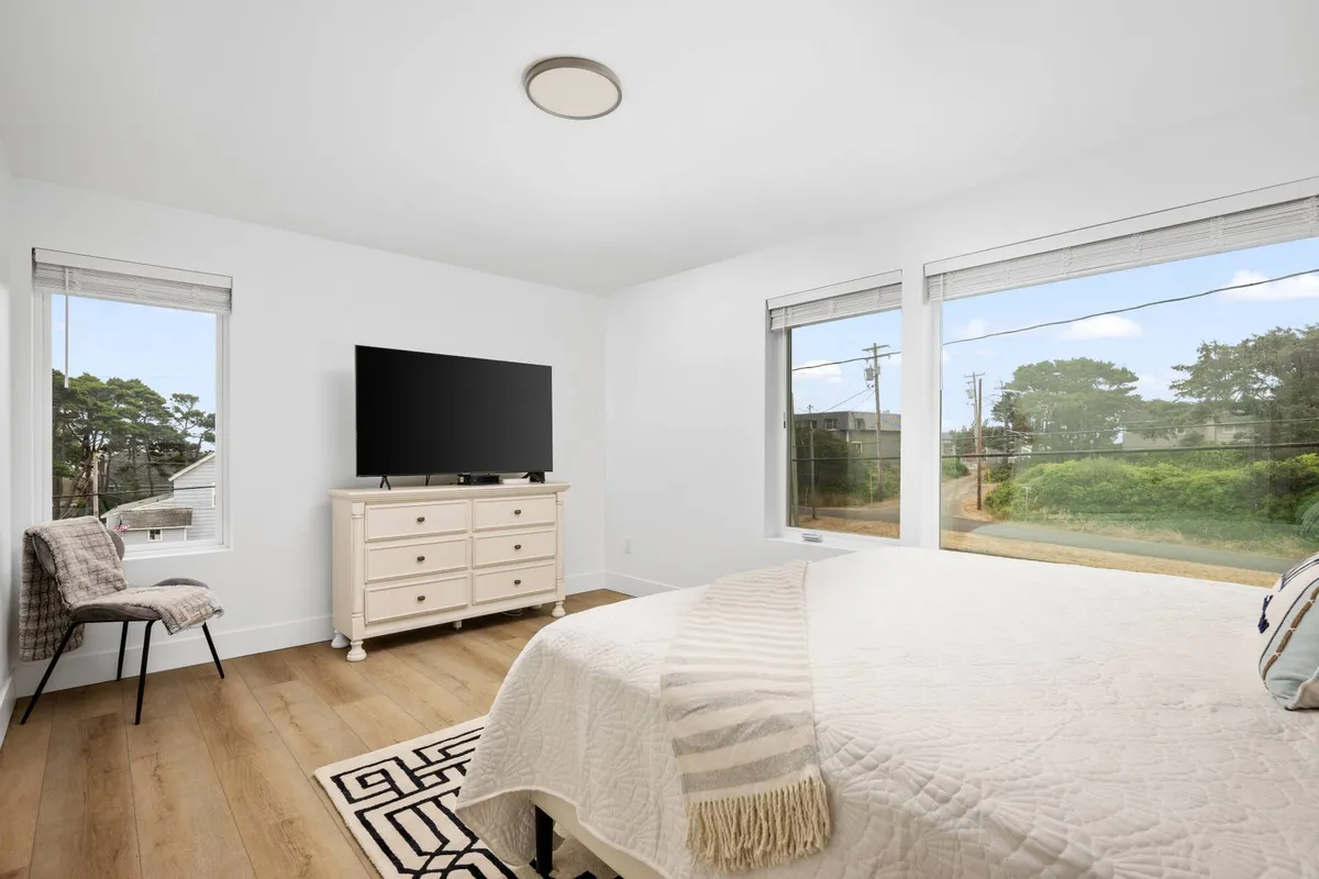 A serene bedroom featuring a spacious bed with plush bedding, a dresser, television, large windows letting in ample natural light, and a cozy chair. The decor is calm and inviting, making this space ideal for relaxation and unwinding after a day of activities.