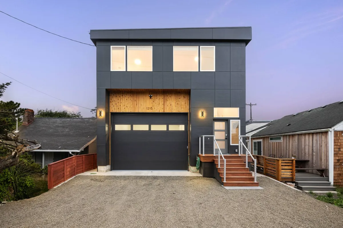 A contemporary home facade featuring a wooden entrance, large windows, and a welcoming stoop with railing. The setting is perfect for gatherings, with the porch bathed in warm evening light and hints of the surrounding neighborhood visible.